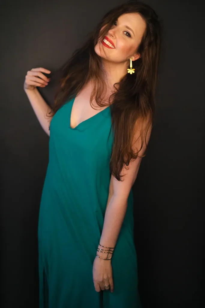 A young woman with long brown hair wearing a green dress smiles broadly. She’s holding a lock of her hair with one hand