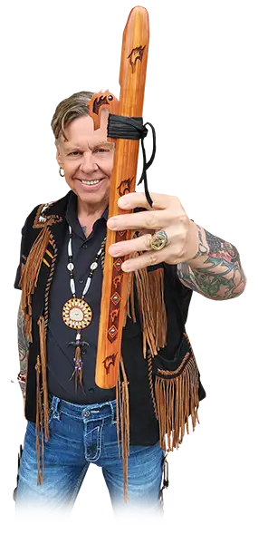 A native man wearing a fringed vest and blue jeans stands smiling at the camera with his hand outstretched, holding a flute.