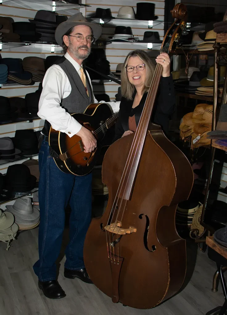 A man and woman standing in a hat shop, the man is holding an acoustic guitar while the woman holds an upright bass.