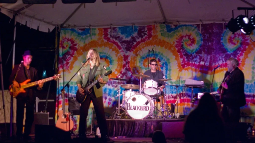 Three guitarists and a drummer performing on a stage at night in front of a tiedye background