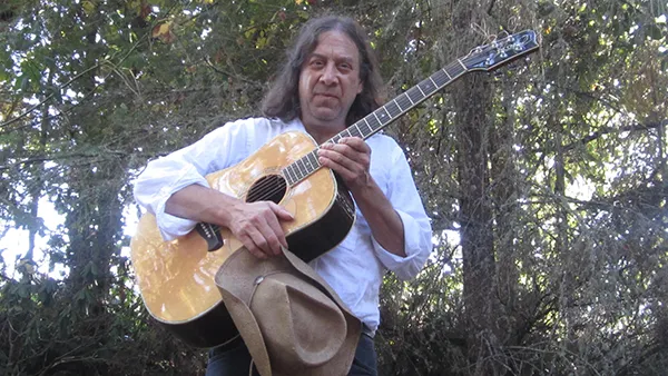 A man in a white dress shirt is standing in the woods holding a guitar, looking down at the camera.
