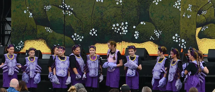 A group of children in matching purple insect costumes standing front of an outdoor stage as an audience looks on from lawn seating
