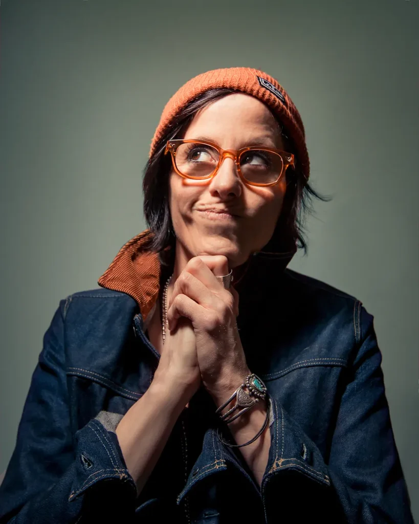 A woman wearing orange eyeglasses and a denim jacket looks upward to the right while smiling from the corner of her mouth.