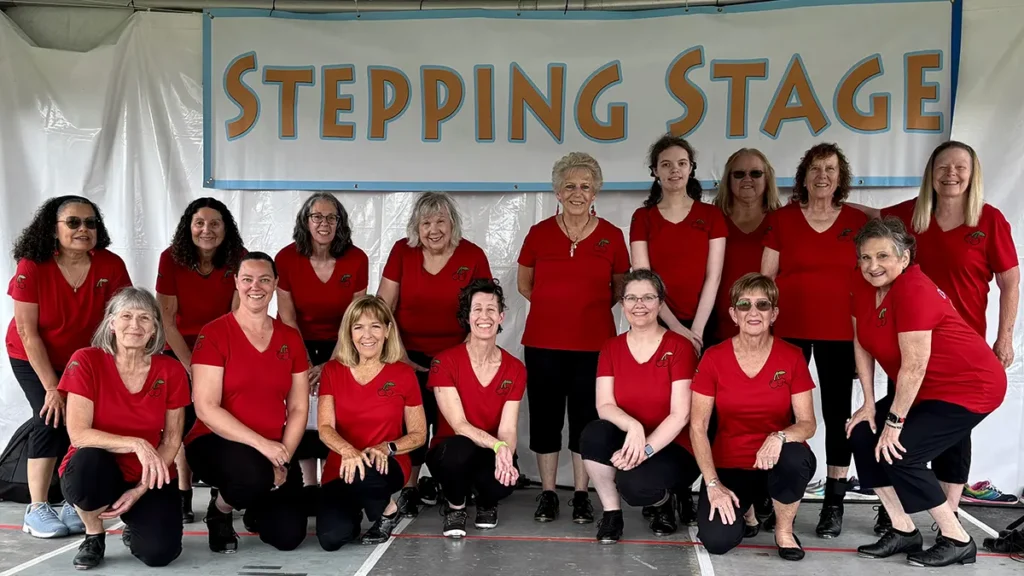 A group of women wearing red t-shirts and black leggings stand and crouch while smiling on a dance stage