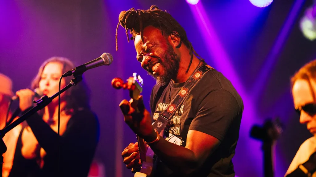 A man with micro dreadlocs tied in a top knot plays guitar on stage, he is backlit in purple light