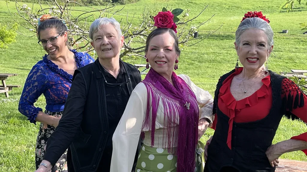 Four women standing smiling in a park, three are wearing ruffled flamenco tops while the fourth is wearing all black