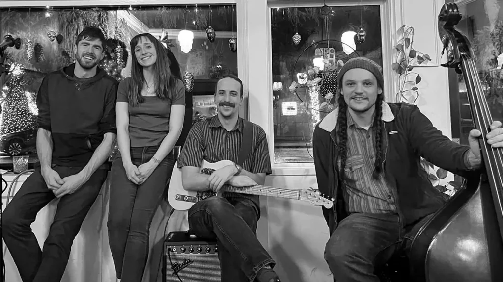 Black and white photo of three men and a woman smiling while sitting in front of a row of shop windows
