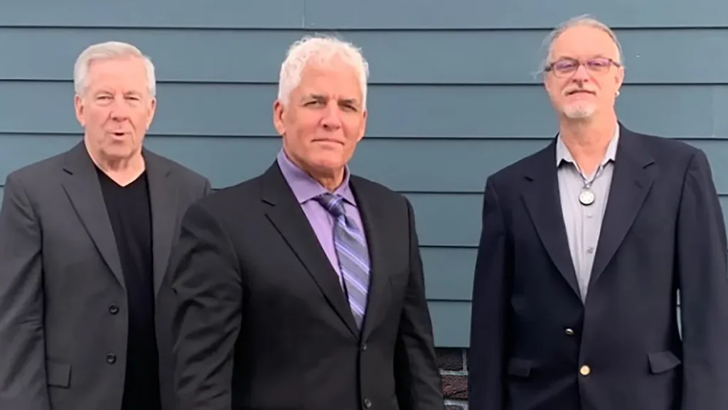 Three men with silver hair wearing suits stand in front of blue siding