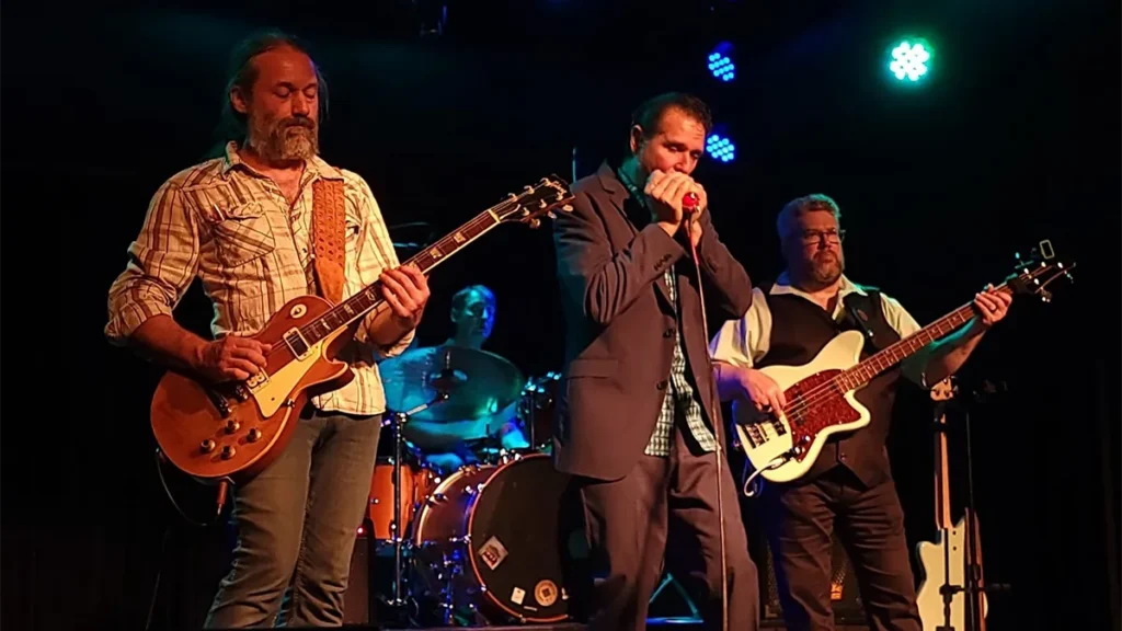 Three men stand on a dark stage. The man on the left is playing an electric guitar, the man in the center is playing harmonica, and the man on the right is playing the bass guitar