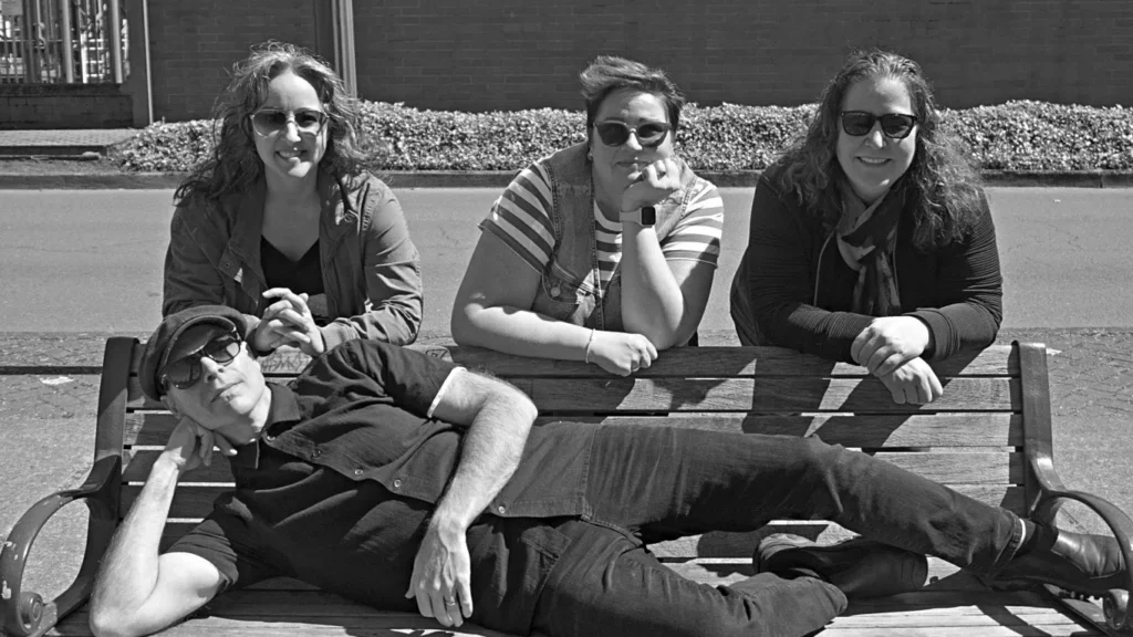 Black and white photo of three women leaning over with their elbows resting on a bus stop bench, a man is laying stretched on the length of the bench