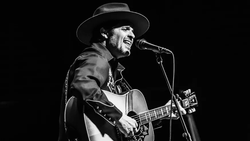 Black and white image of a man in a stetson hat with short facial hair playing guitar standing in front of a microphone