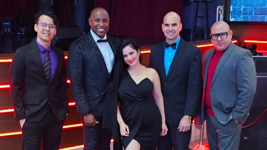 Three men in suits and a woman in a black dress stand in a dance floor in front of a stage