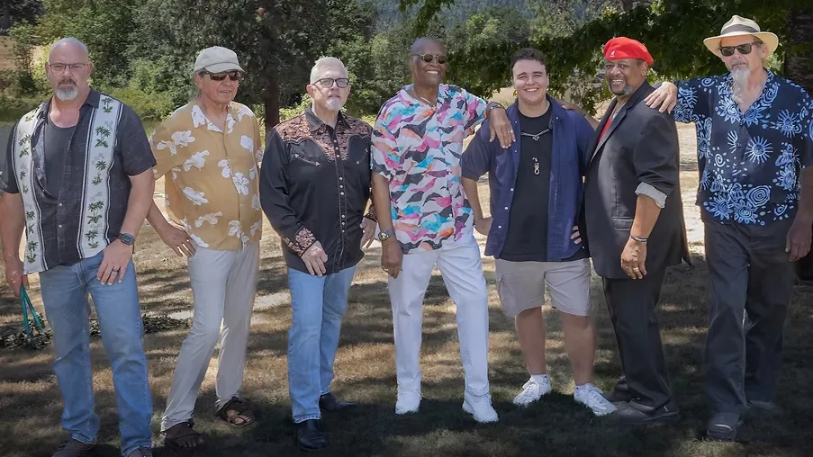 Seven men stand smiling in a row outdoors under the shade of trees