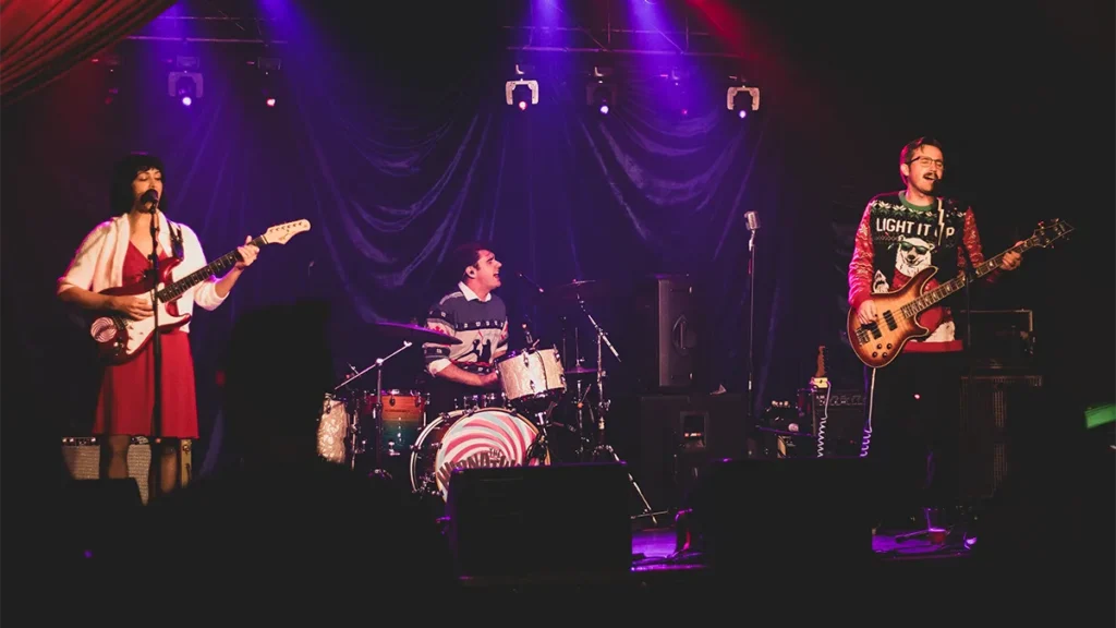 A woman playing guitar, a man playing drums, and a man playing bass sing on a stage