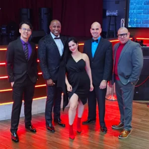 Three men in suits and a woman in a black dress stand in a dance floor in front of a stage