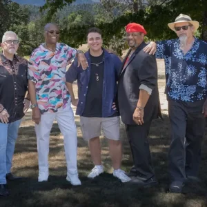 Five men stand smiling in a row outdoors under the shade of trees