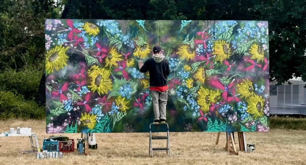 A man stands outdoors on top of a step stool, painting a floral mural on a large canvas.